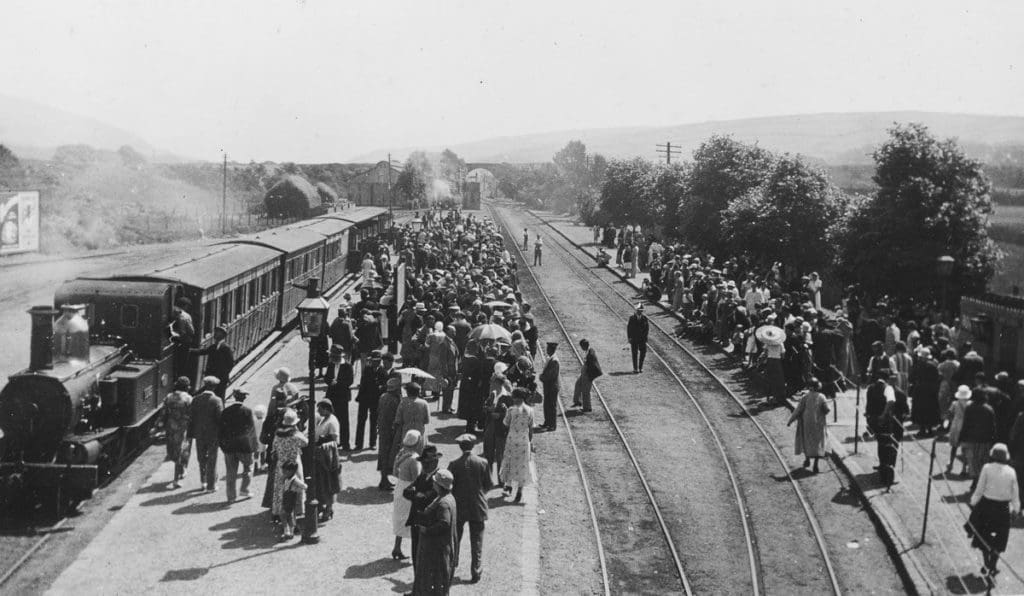 Black and white image of people stood on a station platform and others crossing the tracks, with a train parked